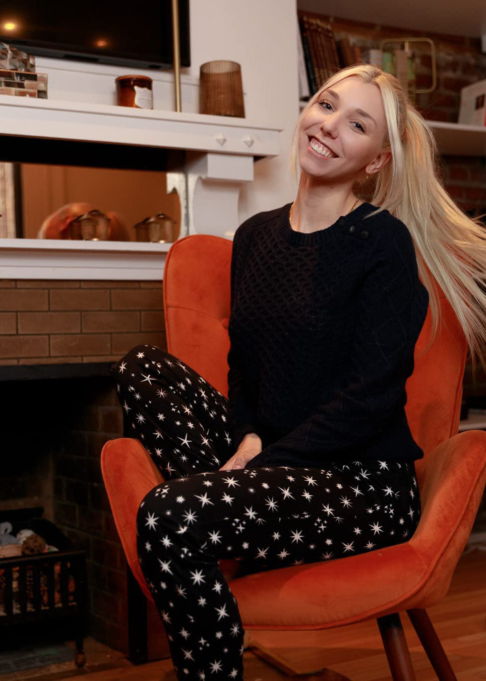 PJ joggers with soft velvety texture, stretch, elastic waistband, drawstring, and stylish ankle cuff. This pattern is scattered stars, of varied size and shape, white, on black stars on a black background. The model is happily sitting on a retro burn orange chair.